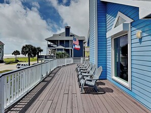 A wooden deck with chairs, a railing, and a building with a blue exterior and an American flag. - Summer Breeze by Ryson Vacation Rentals (Galveston)