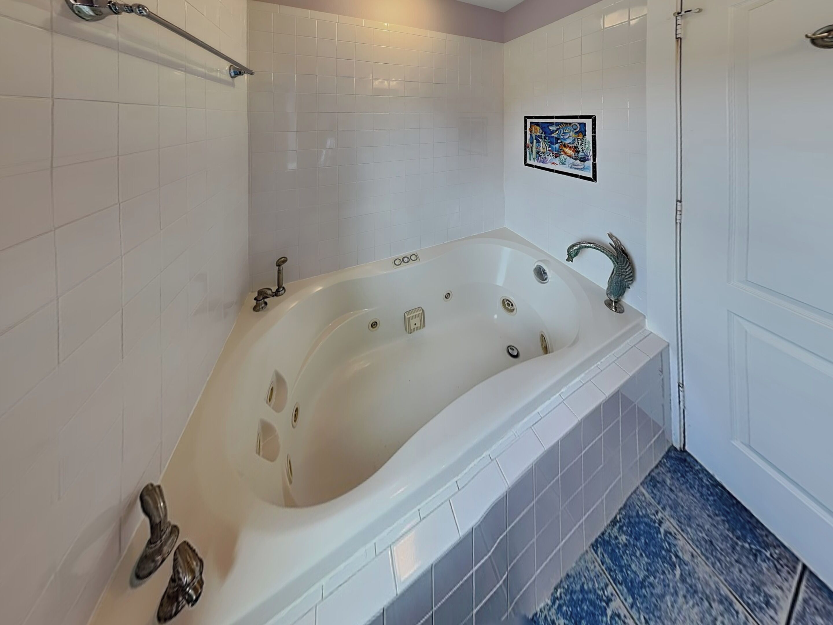 Un baño con una bañera grande, paredes de azulejos blancos, zona de ducha y suelo con estampado azul.