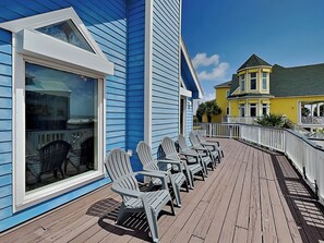 Een houten terras met Adirondack-stoelen, een blauw gebouw met witte details en een geel gebouw op de achtergrond.