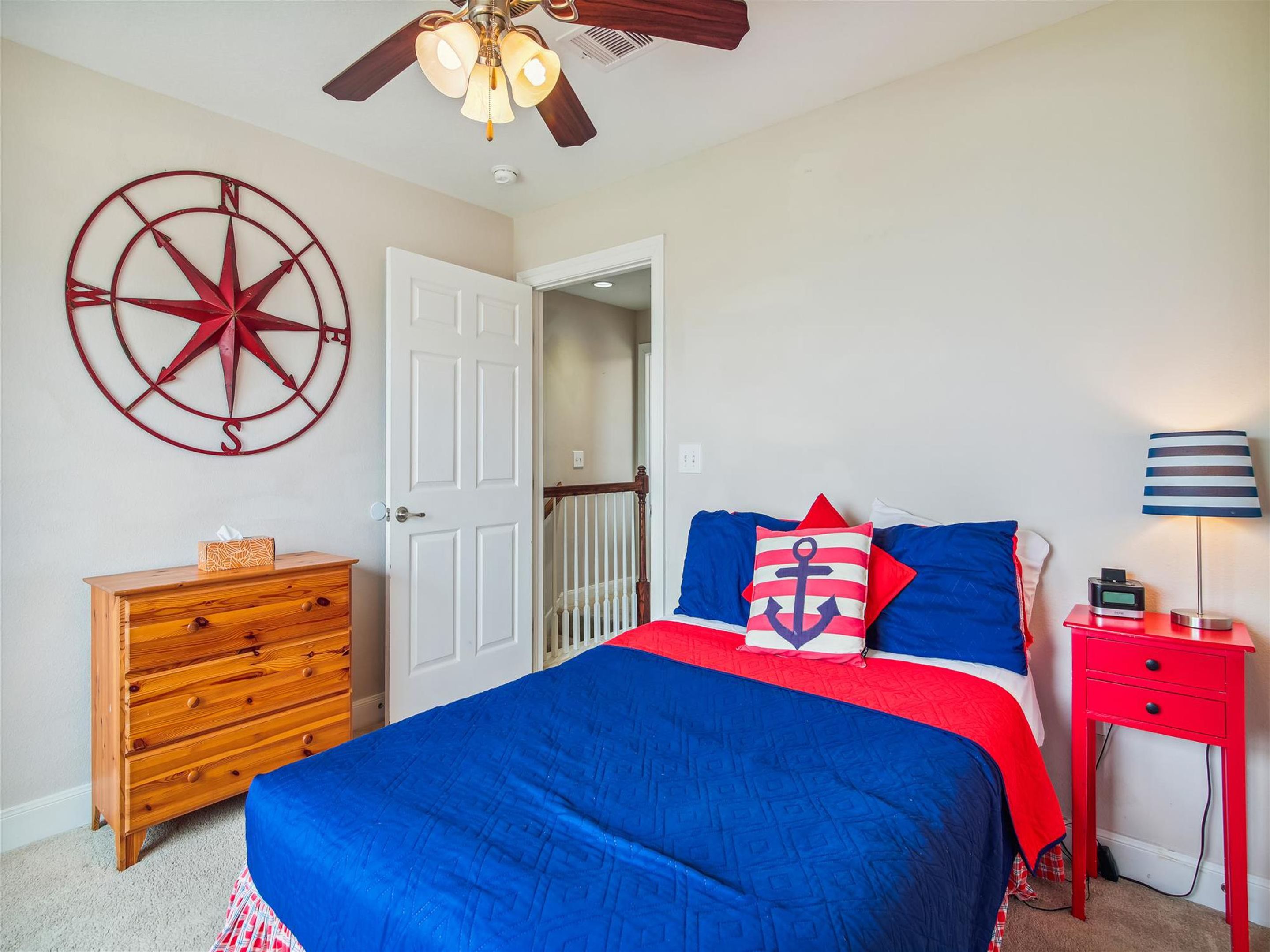 A bedroom with a bed, a wooden dresser, a red nightstand, a wall-mounted compass, and a ceiling fan.