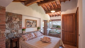 Un dormitorio con pared de piedra, cama con ropa de cama floral, techo de madera y vista al exterior a través de una ventana.