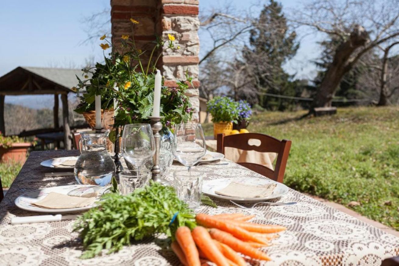 An outdoor dining table set with plates, glasses, a pitcher, and fresh vegetables.