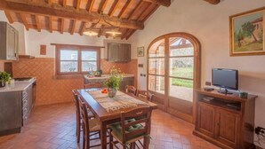 Cocina rústica con vigas de madera, una mesa de comedor con sillas, un televisor y un ventanal con vistas al exterior.