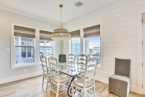 A dining area with a glass table surrounded by chairs, a hanging light fixture, and large windows with blinds.