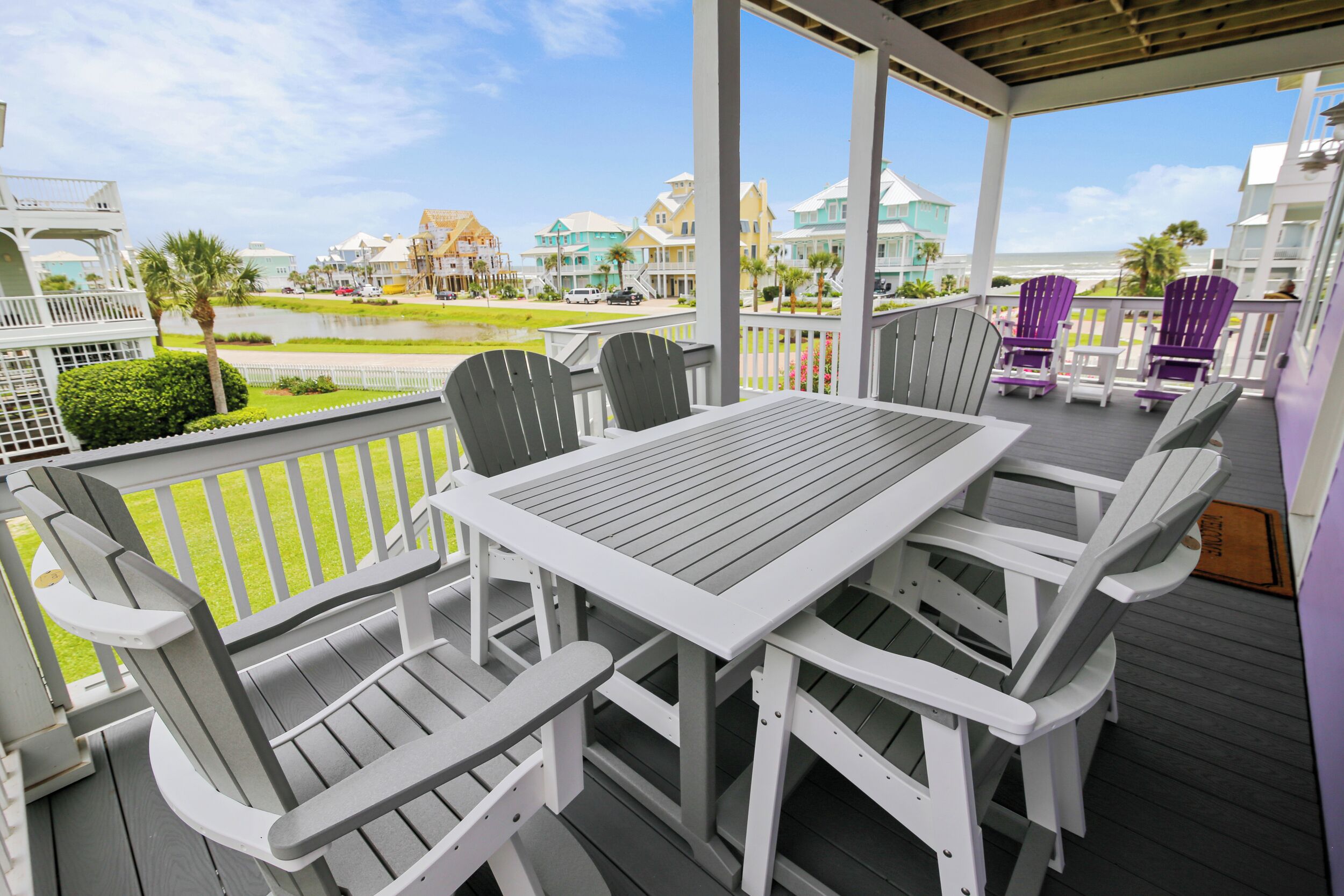 A covered outdoor seating area with a table and chairs, overlooking a residential area with houses and a body of water.