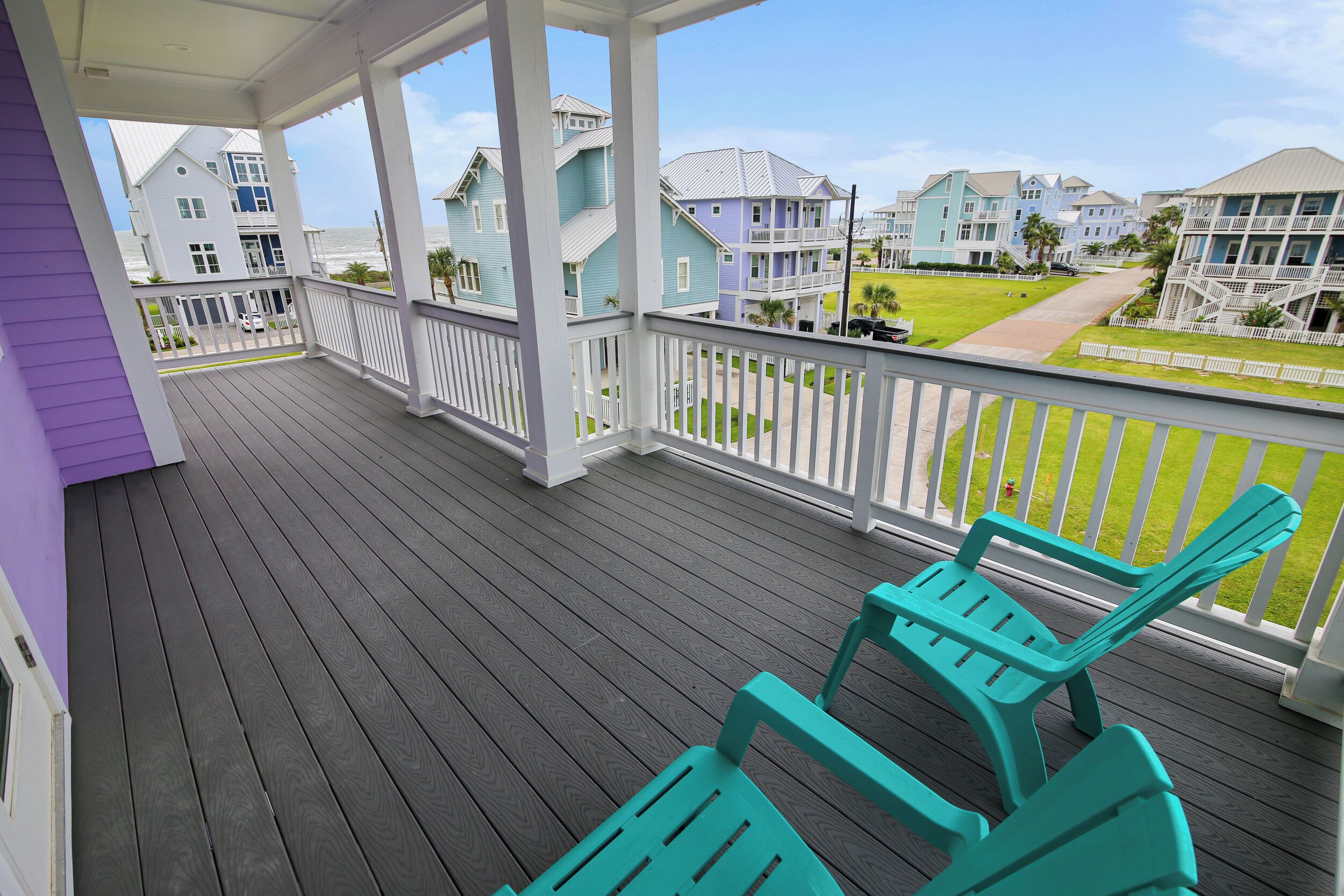 Um deck de madeira com cadeiras cor turquesa, com vista para uma área residencial com casas e gramado.