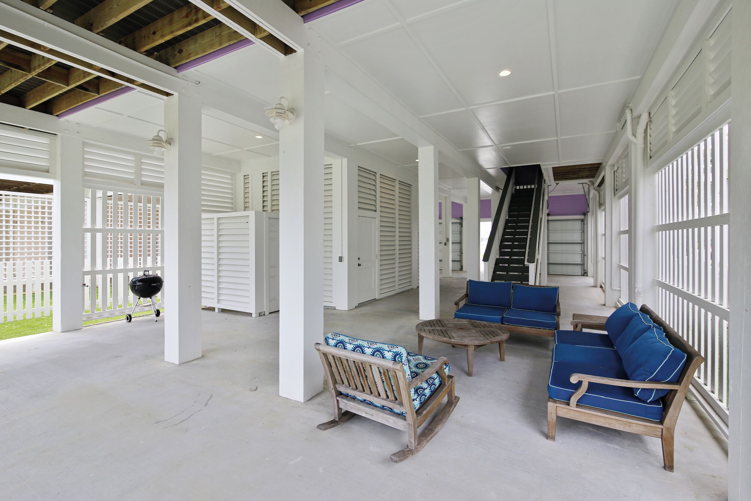 A covered outdoor area with white columns, blue and wooden furniture, and a staircase leading to an upper level.