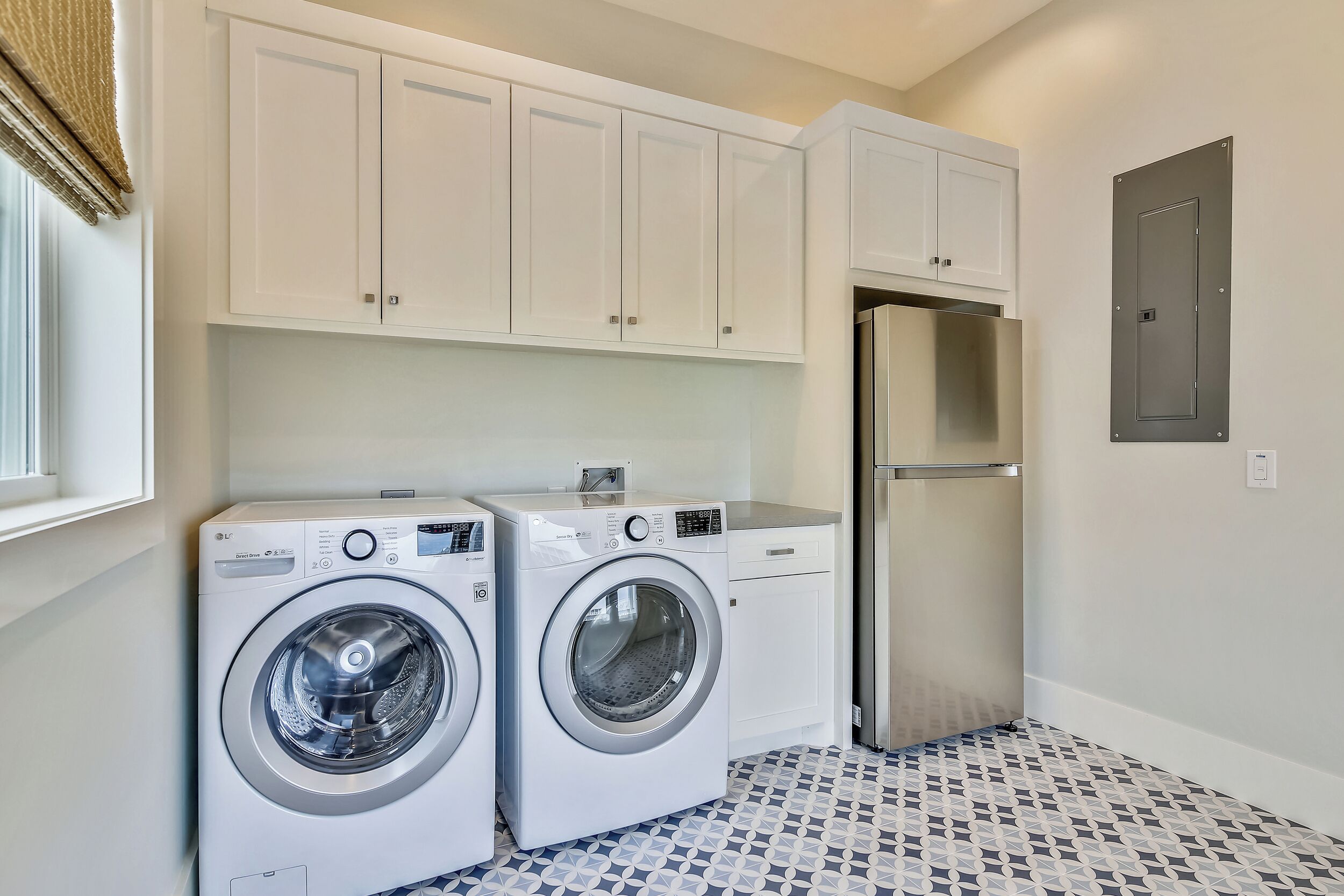 A laundry room with two front-loading washing machines, a stainless steel refrigerator, and white cabinets.