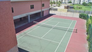 An outdoor tennis court with a net, surrounded by a red and green surface, adjacent to a building with a red exterior and a covered area. - Room with a View by Ryson Vacation Rentals (Galveston)