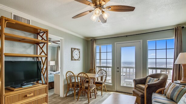 A spacious living room with a television, wooden furniture, a ceiling fan, and large windows offering a view of the ocean. - Room with a View by Ryson Vacation Rentals (Galveston)