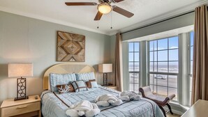 A bedroom with a large bed, two bedside tables, a ceiling fan, a view of the ocean, and a window with curtains. - Room with a View by Ryson Vacation Rentals (Galveston)