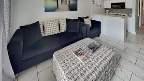 A modern living room with a dark sofa, a woven ottoman, and a kitchen area with white cabinets and a microwave. - All for Fun by Ryson Vacation Rentals (Galveston)