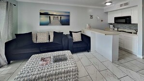 A modern living room with a dark sofa, patterned ottoman, and a painting of a pier. A kitchen with white cabinets and a microwave is visible. - All for Fun by Ryson Vacation Rentals (Galveston)