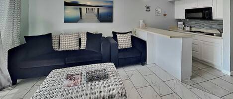 A modern living room with a dark sofa, patterned ottoman, and a painting of a pier. A kitchen with white cabinets and a microwave is visible.