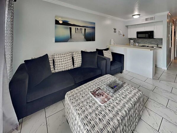 A modern living room with a dark sofa, a woven ottoman, and a kitchen area with white cabinets and a microwave. - All for Fun by Ryson Vacation Rentals (Galveston)