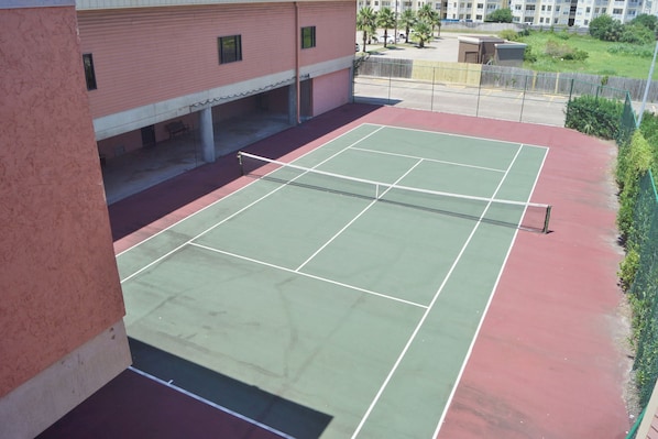 An outdoor tennis court with a net, surrounded by a building with a red exterior and a covered seating area.