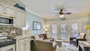A kitchen with a stove, microwave, and cabinets, a beige sofa with pillows, a ceiling fan, and a window with blinds. - Ocean Oasis by Ryson Vacation Rentals (Galveston)