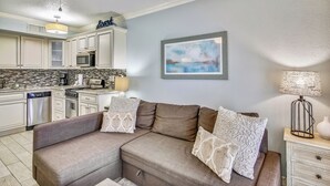 A modern kitchen with white cabinets, a microwave, and a dishwasher. There is a beige sofa with patterned cushions in the living area. - Ocean Oasis by Ryson Vacation Rentals (Galveston)