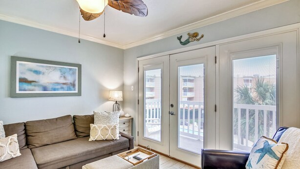 A living room with a brown sectional sofa, a small ottoman, a dark leather armchair, a ceiling fan, and a large window with blinds. - Ocean Oasis by Ryson Vacation Rentals (Galveston)