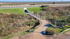 A pedestrian bridge over a road with a view of the ocean in the background. - Wine Down by Ryson Vacation Rentals (Galveston)