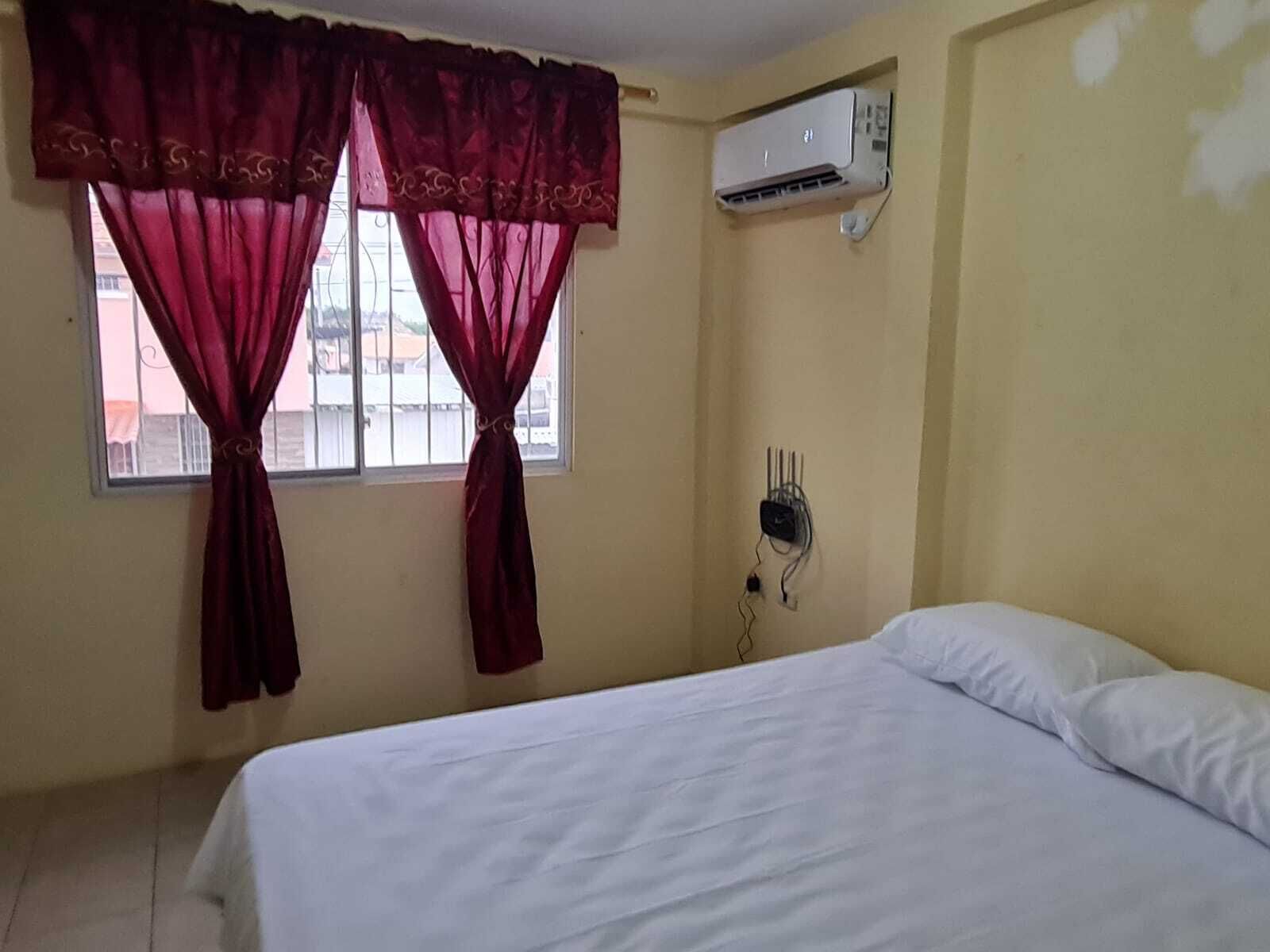 A bedroom with a bed, a window with curtains, an air conditioning unit, and a wall socket.