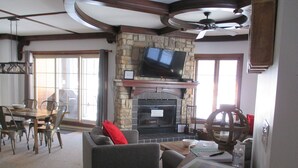 A living room with a stone fireplace, a flat-screen TV mounted above it, a dining area with a table and chairs, and a ceiling fan.
