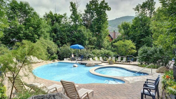 A swimming pool with lounge chairs and a blue umbrella, surrounded by trees and a brick patio.