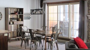A dining area with a wooden table and chairs, a kitchen area with cabinets and shelves, and a large window with curtains.