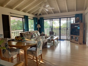 A spacious living area with a wooden ceiling, a ceiling fan, a large window with curtains, a sofa, a dining table with chairs, and a television on a wooden cabinet. - SPACIOUS 2BD/2BA PRIVATE QUIET W/ CORNER LANAI IN MOLOKAI (Maunaloa)