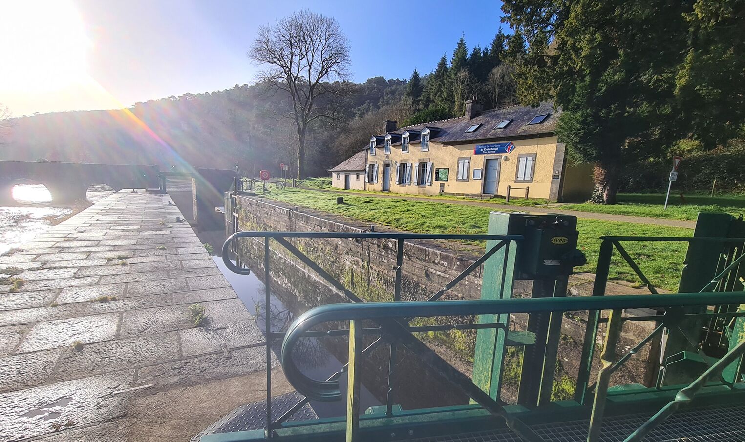 A riverside walkway with a lock gate, a small building with a sign, and a clear sky.