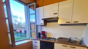 Cocina compacta con armarios blancos, estufa a gas, cafetera y ventana con vistas a un área verde.