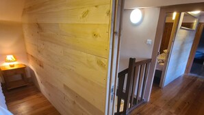 Habitación con paneles de madera, una cama, una mesita de noche y una lámpara. Se ve una barandilla de madera y una puerta que da a otra habitación.