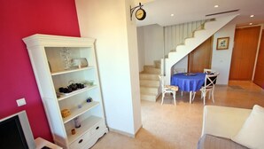 Una sala de estar con un sofá blanco, un mueble para televisión, una estantería, una mesa de comedor con sillas y una escalera.