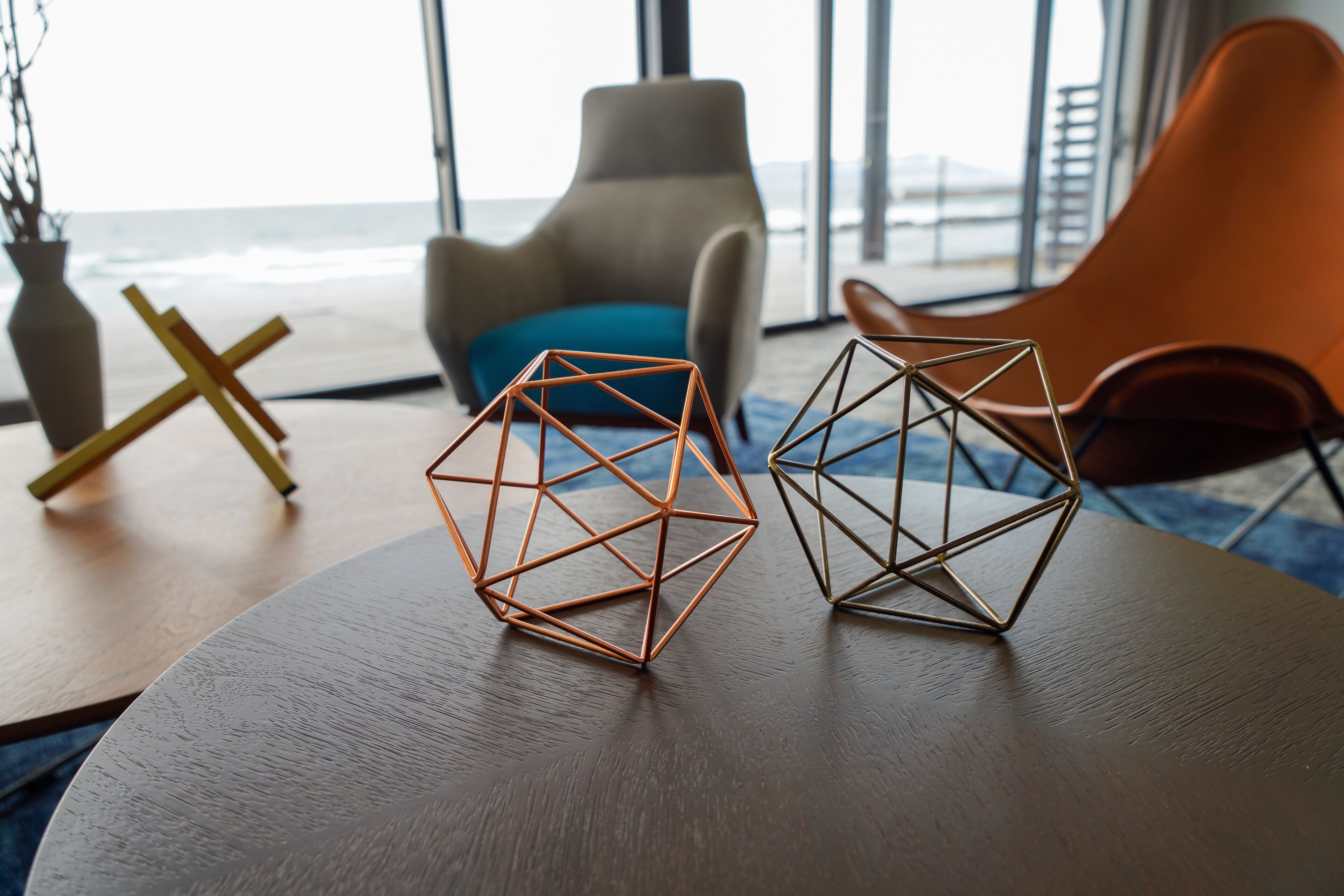A modern room with a view of the ocean, featuring two geometric metal sculptures on a wooden table, a beige armchair, and an orange chair.