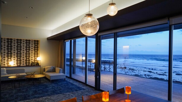 Una sala de estar moderna con un ventanal grande con vistas a la playa, una mesa de comedor de madera con velas y un papel tapiz decorativo.