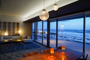Una sala de estar moderna con un ventanal grande con vistas a la playa, una mesa de comedor de madera con velas y un papel tapiz decorativo.