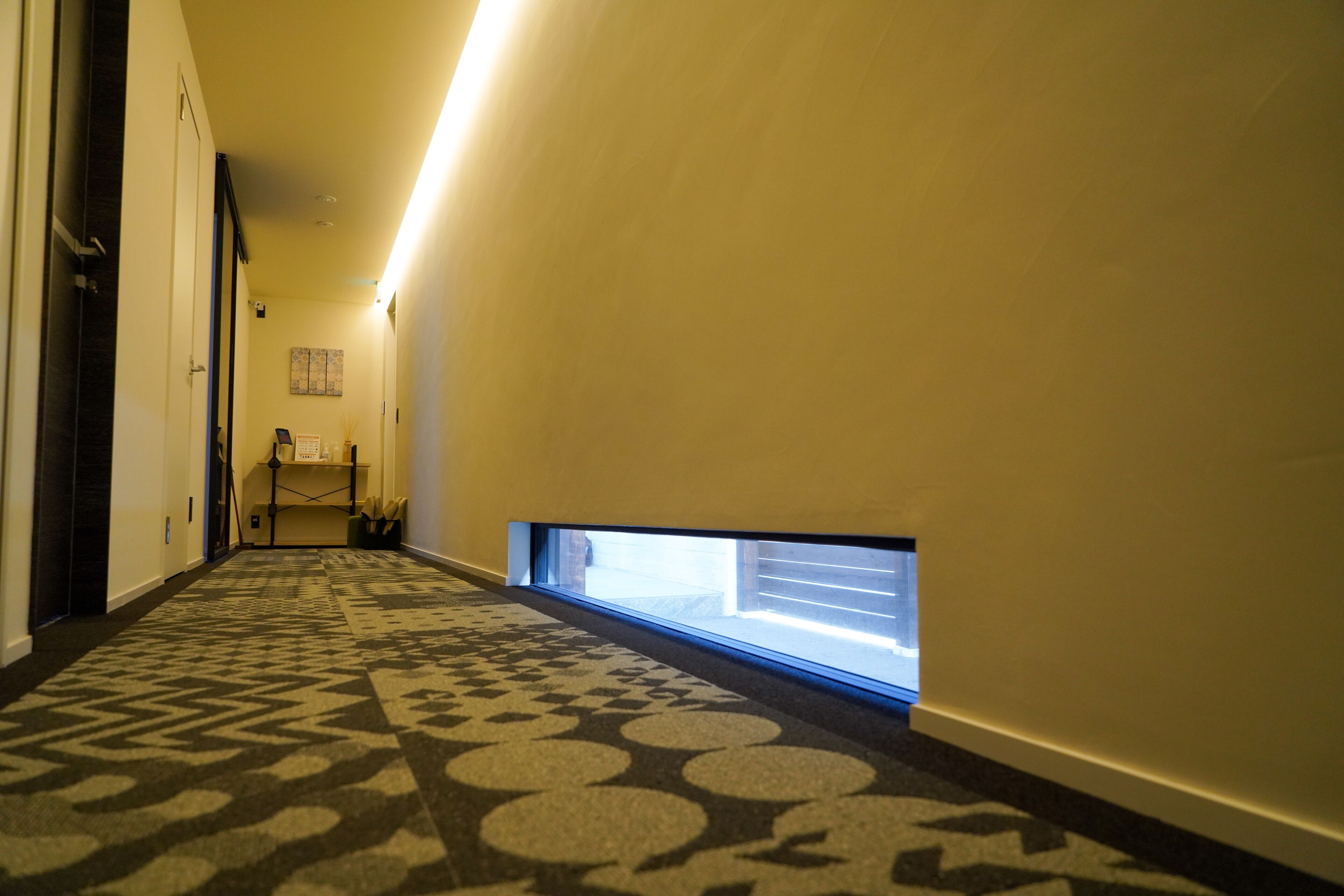 A hotel hallway with patterned carpet, a small window, and a door on the left.