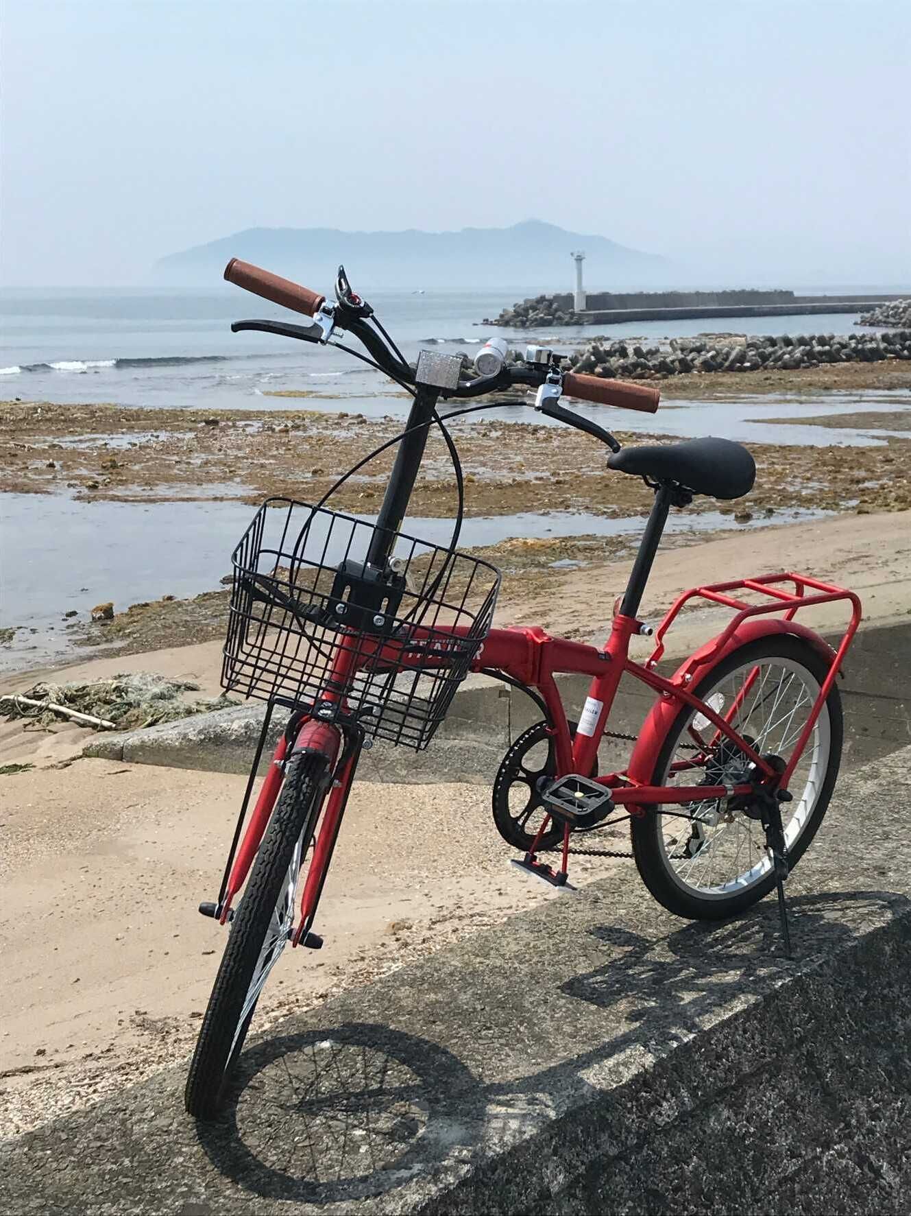 A red bicycle with a basket and a seat, parked on a concrete surface near a beach with a rocky shoreline and a distant mountain.