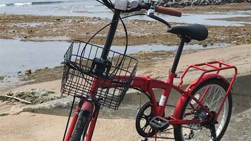 A red bicycle with a basket and a seat, parked on a concrete surface near a beach with a rocky shoreline and a distant mountain.