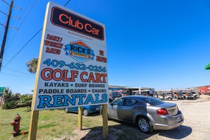 Une affiche indiquant les tarifs journaliers et hebdomadaires de location de véhicules Club Car, ainsi que le numéro de téléphone à contacter et le matériel disponible, comme des voiturettes de golf, des kayaks, des planches de surf, des planches de paddle et des chaises.