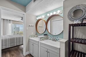 Une salle de bain avec deux lavabos, un meuble-lavabo et un grand miroir avec éclairage. Il y a une fenêtre avec des rideaux et une vue sur un bâtiment à l'extérieur.