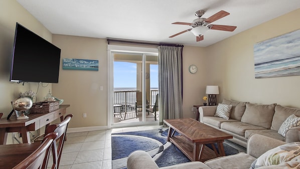 A living room with a TV, a ceiling fan, a sofa, a coffee table, and a view of the beach through a sliding door.