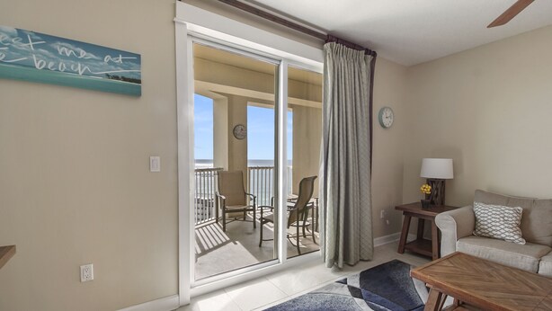 A room with a sliding glass door leading to a balcony with chairs, a sofa, a lamp, and a clock on the wall. - Grand Panama Resort beautiful condo (Panama City Beach)
