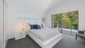 A modern bedroom with a large bed, a nightstand, a desk, and a chair. There is a ceiling fan and a large window with a view of trees. - Laze @ Lighthouse - modern family home with views and pool (Port Macquarie)