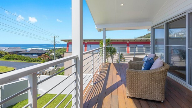 A balcony with a wicker sofa and cushions, overlooking a coastal area with buildings and a clear sky. - Laze @ Lighthouse - modern family home with views and pool (Port Macquarie)
