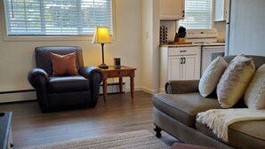 A living room with a sofa, armchair, and ottoman, a small table with a lamp, and a kitchen area visible through an open doorway.
