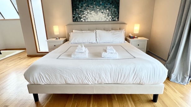 A large bed with white linens, two bedside tables with lamps, a large abstract painting above the bed, and a view of the outside through a window. - 💫🏡6 bedroom/3.5 bath home private acreage parkings (Mission)
