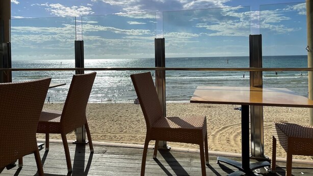Un restaurante frente al mar con sillas de mimbre y barandilla de vidrio con vistas al océano.