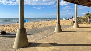Una playa con una zona cubierta sostenida por pilares de hormigón, arena y el océano al fondo.