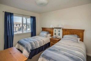 A hotel room with two beds, a wooden headboard, a nightstand with lamps, a small table, and a window with curtains.